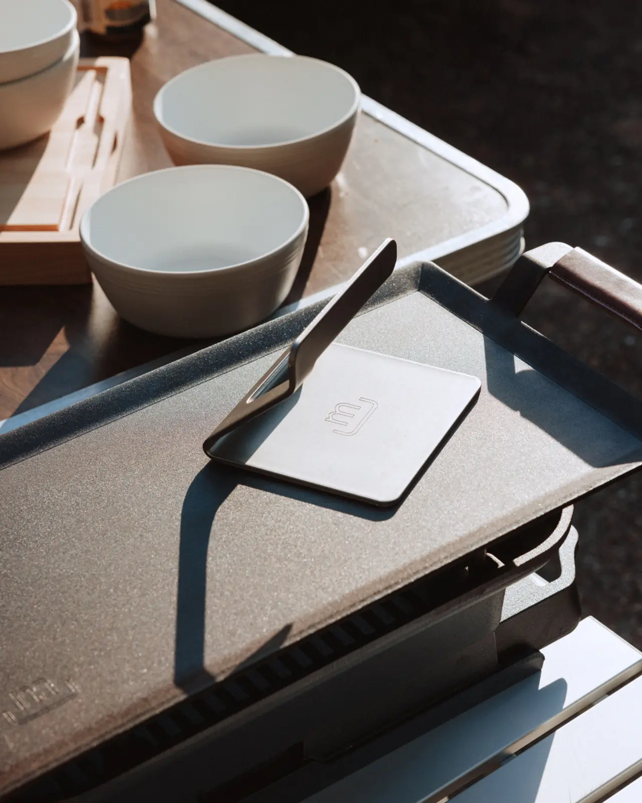 Sunlight casts shadows on a kitchen countertop with bowls, utensils, and a portable cooking appliance.