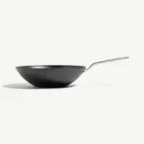 A black, round wok with a long handle sits on a light background.