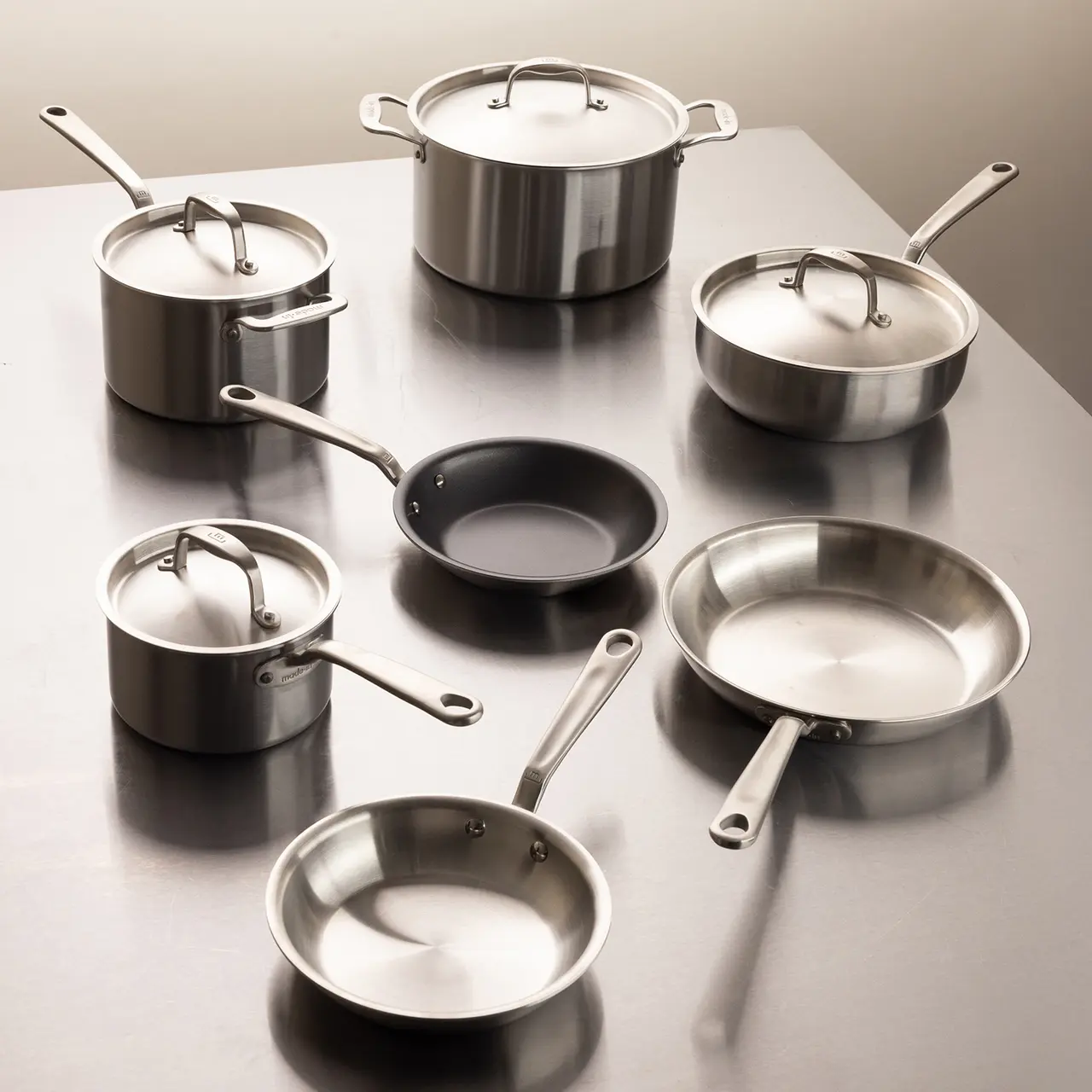 A variety of stainless steel pots and pans with lids on a metallic surface angled to show their different sizes and shapes.