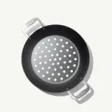 A round, perforated cooking pan with two metallic handles rests on a light background.