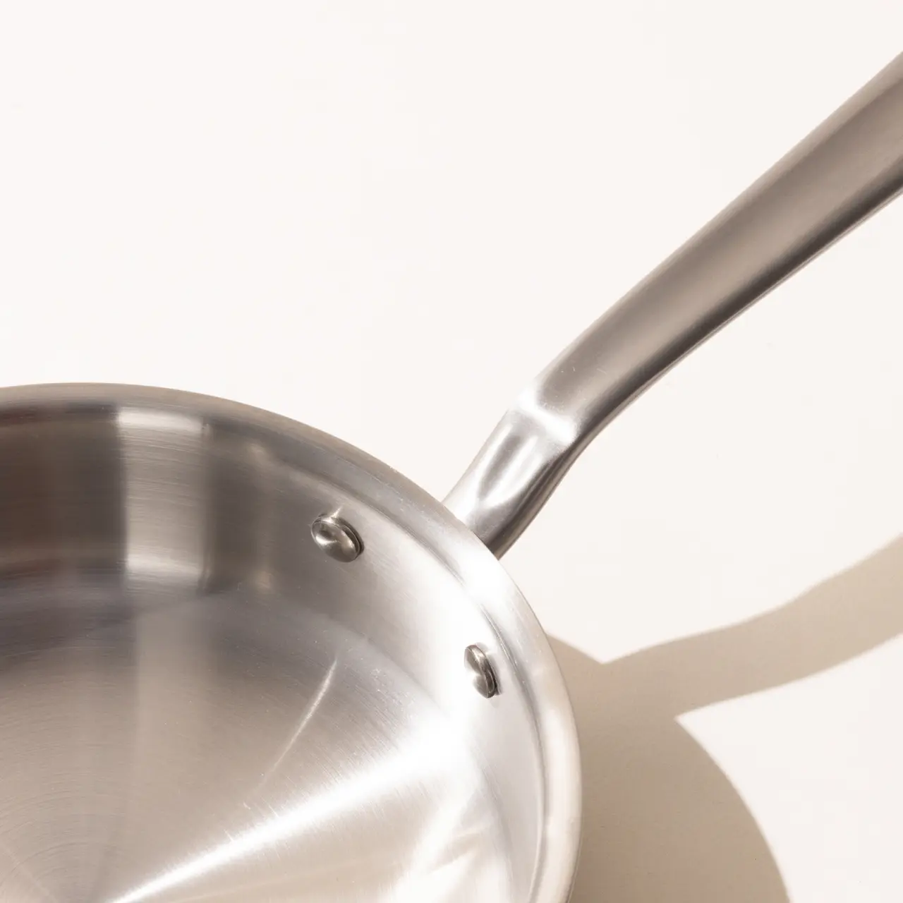 A stainless steel frying pan with a long handle on a light background, casting a soft shadow.