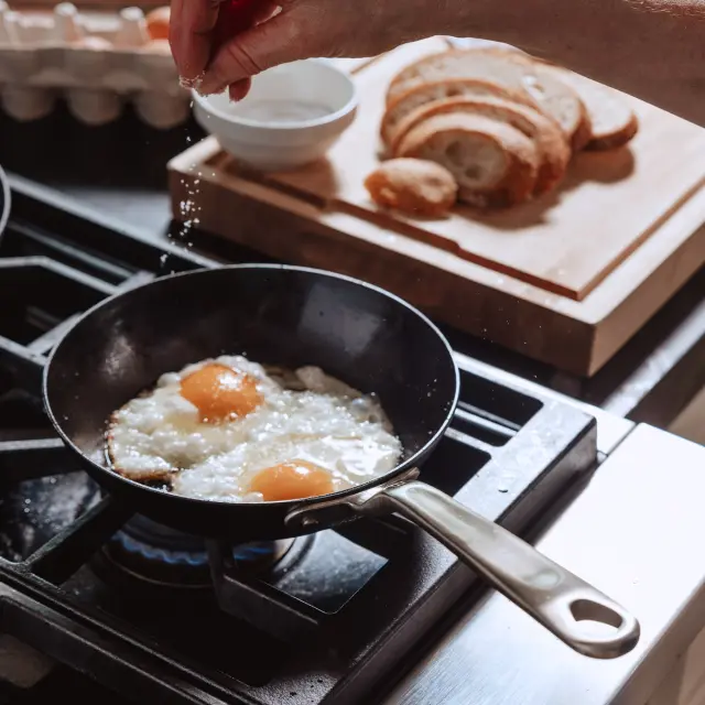 A hand sprinkles salt over two frying eggs in a skillet on the stove, with a bowl and sliced bread in the background.