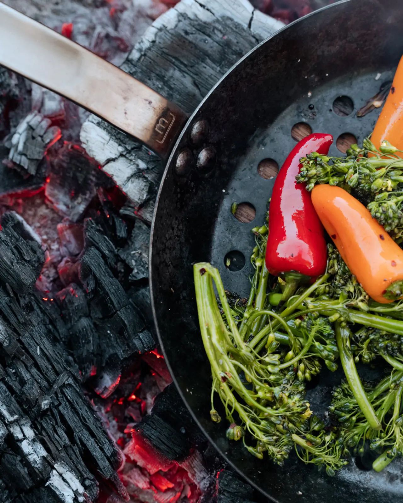 A skillet with vegetables is being cooked over red-hot charcoal embers.
