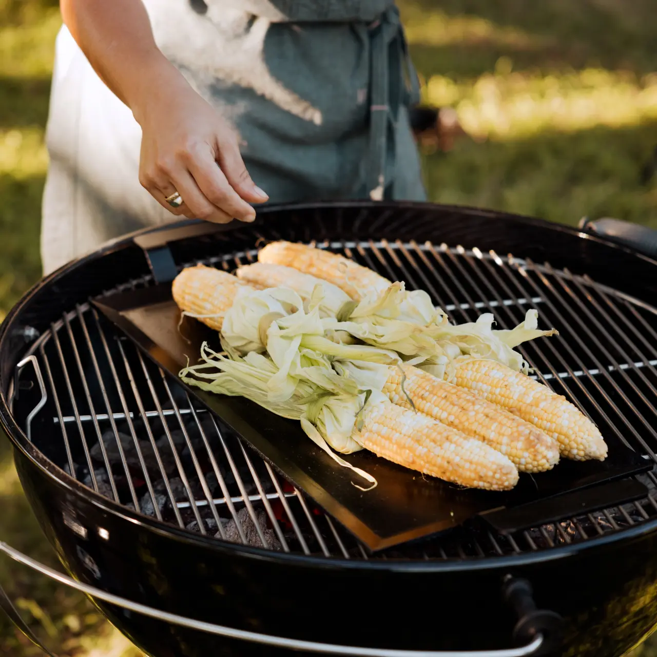 A person seasons corn on the cob on a charcoal grill outdoors.