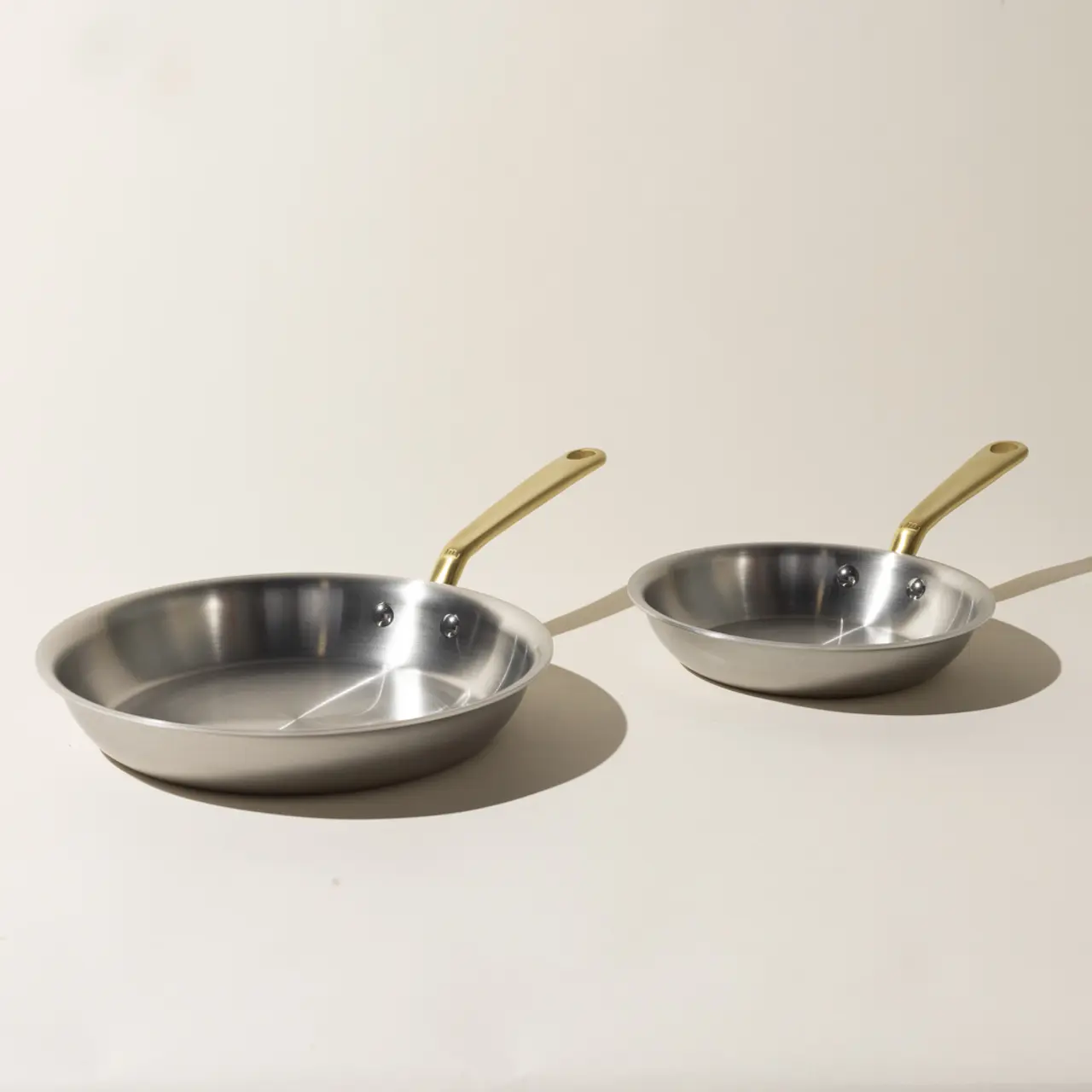 Two stainless steel frying pans with golden handles are placed against a light background.