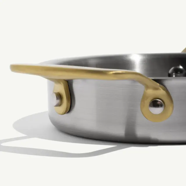 A stainless steel pan with a golden handle rests on a flat surface, casting a soft shadow.