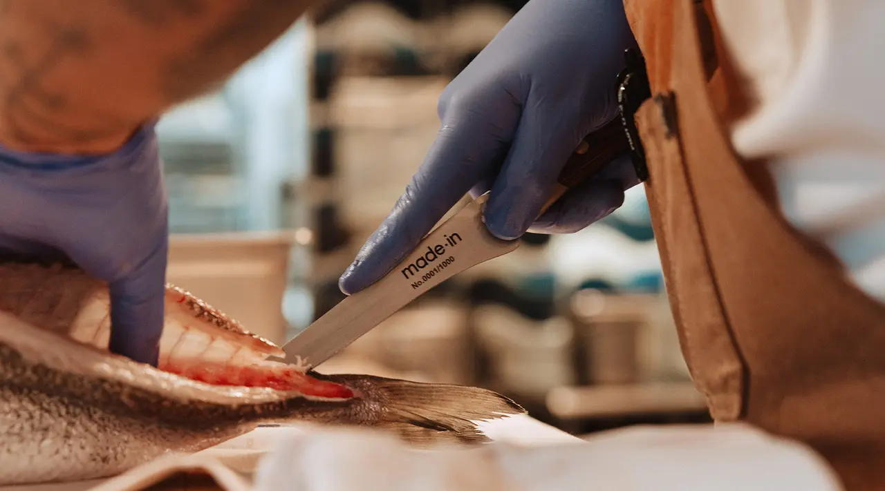 A person wearing gloves is filleting a fish with a knife that has "made in" written on the blade.