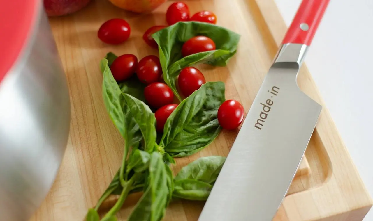 A sharp kitchen knife with a red handle is displayed on a wooden cutting board alongside fresh basil leaves and cherry tomatoes, ready for meal prep.
