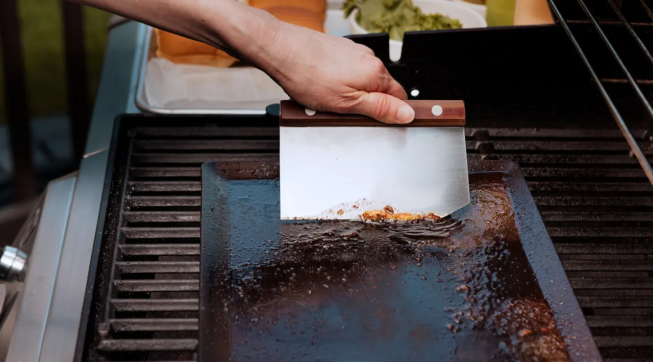 A person's hand cleans a griddle with a scraper on an outdoor grill.
