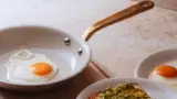 A white frying pan with a golden handle contains a sunny-side-up egg, accompanied by toasted bread topped with avocado and another egg.