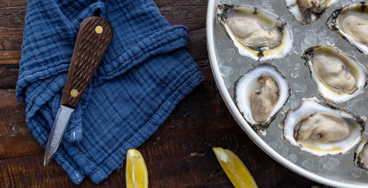 A platter of fresh oysters on the half shell with a shucking knife and lemon wedges on a wooden table with a blue cloth.