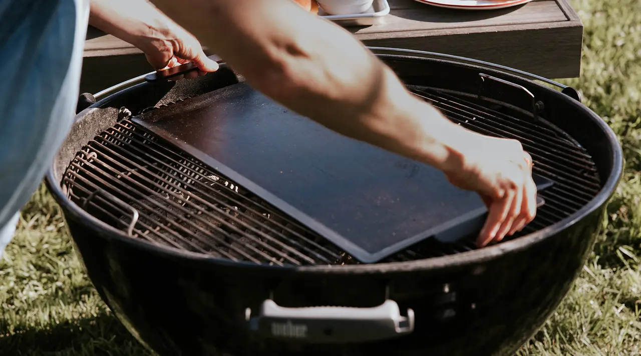 Hands prepare a barbecue grill with a clean flat plate for cooking outdoors.
