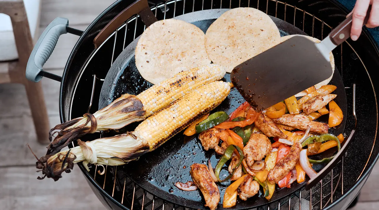 A variety of grilled foods including corn on the cob, tortillas, and mixed vegetables are being cooked on a barbecue grill with a spatula in hand.