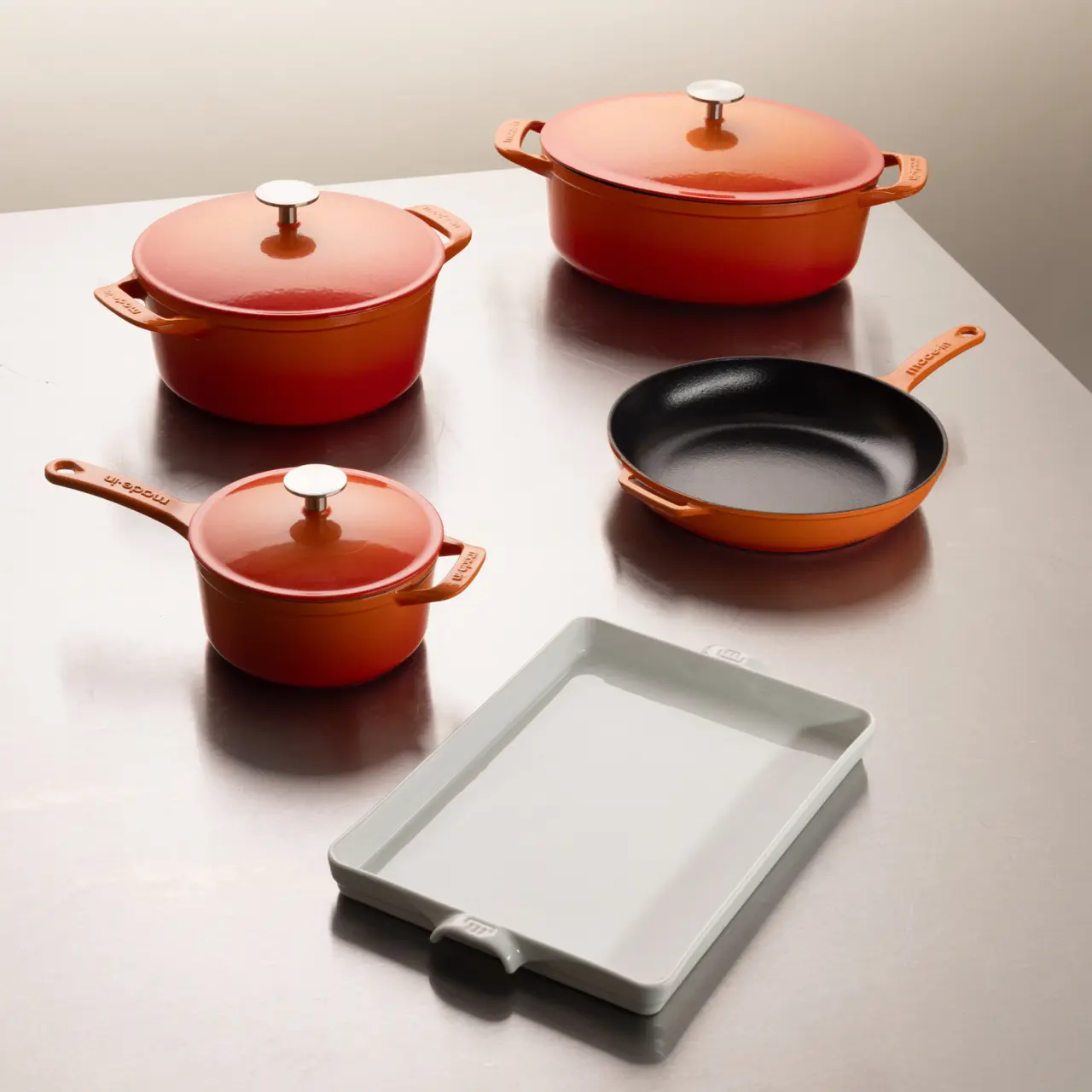 A collection of orange cookware, including various pots with lids and a frying pan, is neatly arranged beside a white cutting board on a tabletop.