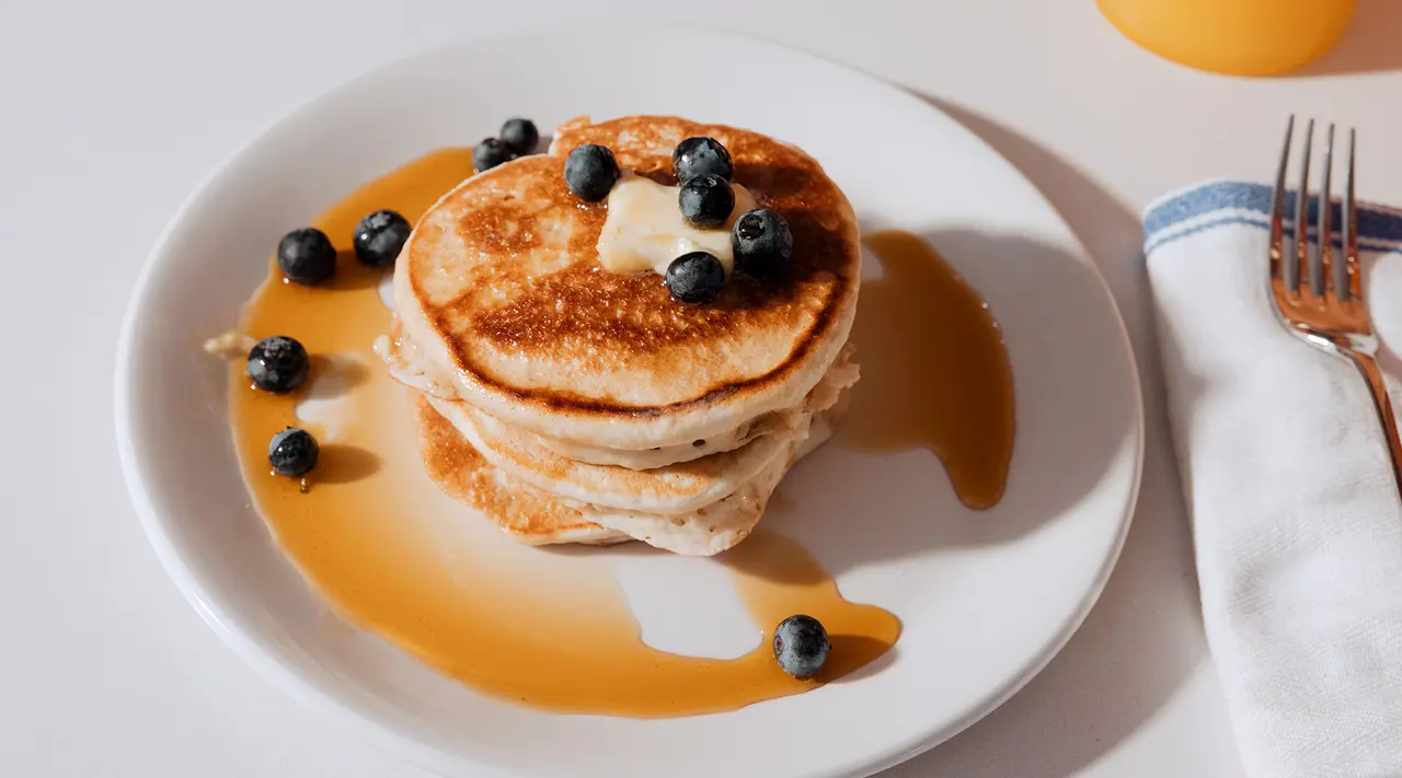 A stack of pancakes topped with butter and blueberries, drizzled with syrup on a white plate, next to a fork and a white napkin.