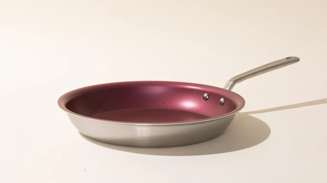 A stainless steel frying pan with a maroon-colored interior sitting on a neutral background with shadows indicating a light source to the left.