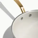 A stainless steel pot with a gold handle features a smooth, white interior and two pouring spouts.