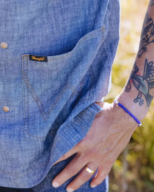 A close-up of a person's hand resting on a denim shirt, showcasing a pocket with a label and a tattoo on their arm.