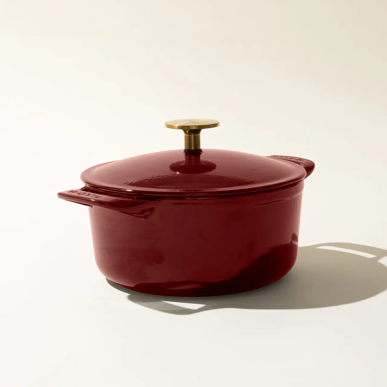 A red enameled cast iron Dutch oven with a lid and a gold-colored knob handle, on a neutral background with a distinct shadow to the right.