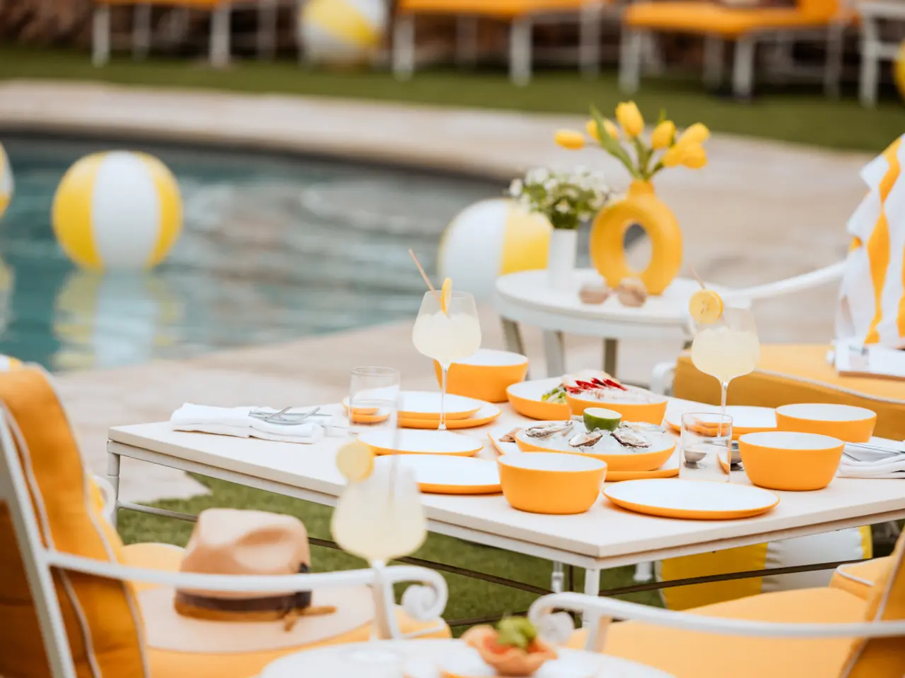 A summer-themed outdoor dining table is set with yellow and white dinnerware, fresh lemons, refreshments, and a straw hat, with a pool and beach balls in the background.