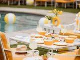 A summer-themed outdoor dining table is set with yellow and white dinnerware, fresh lemons, refreshments, and a straw hat, with a pool and beach balls in the background.