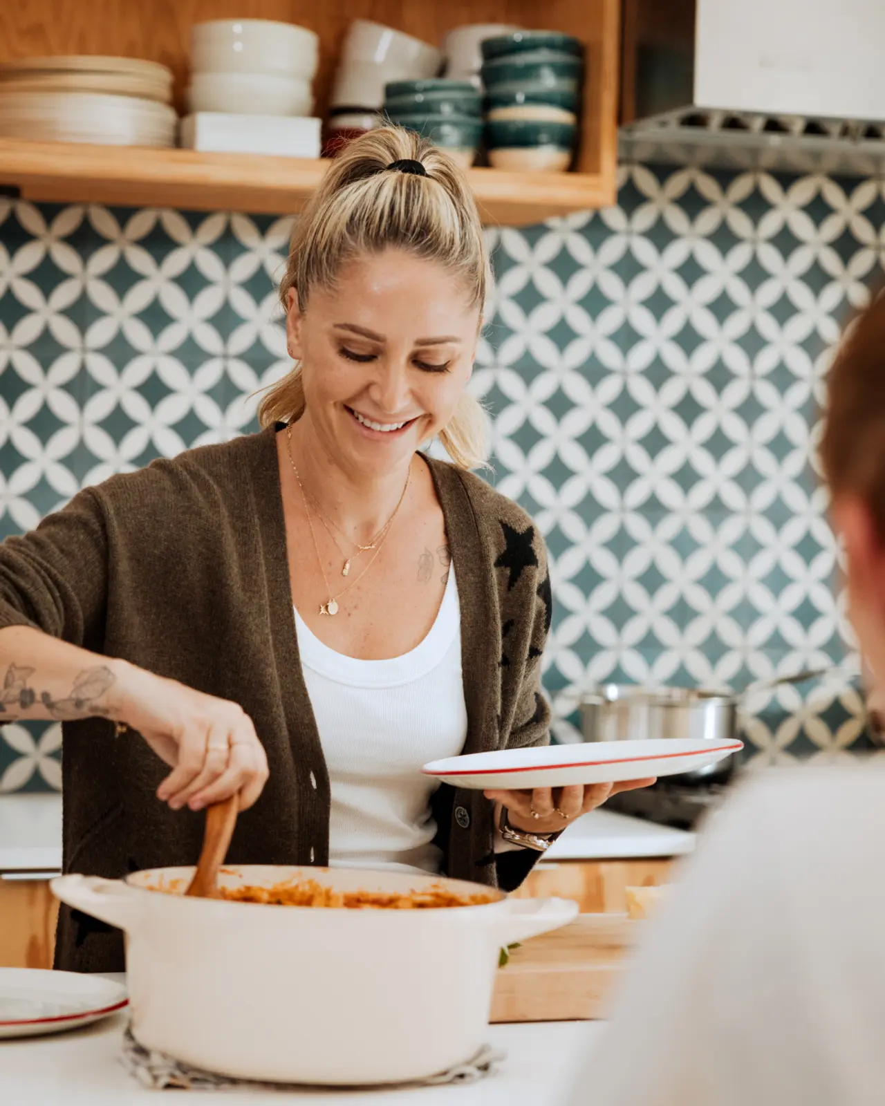 A smiling person is stirring a pot of food while another individual waits with an empty plate.
