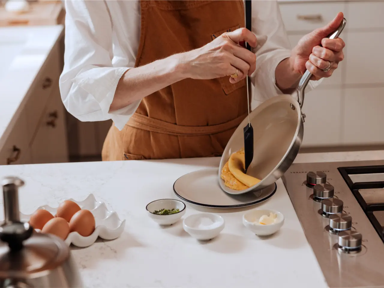 A person in a kitchen is transferring an omelette from a pan to a plate, with various ingredients and eggs in bowls on the counter.