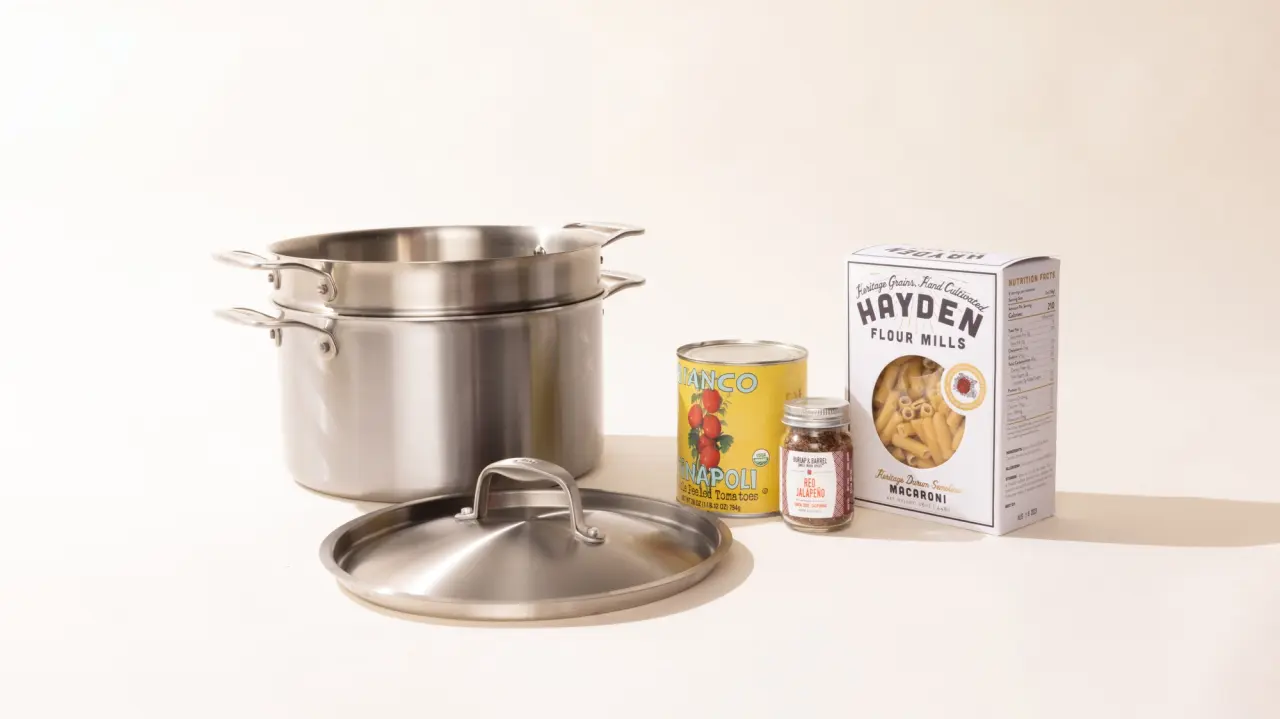 A stainless steel pot with its lid beside it, flanked by a can of tomatoes, a small jar of seasoning, and a box of Hayden Flour Mill pasta on a plain background.