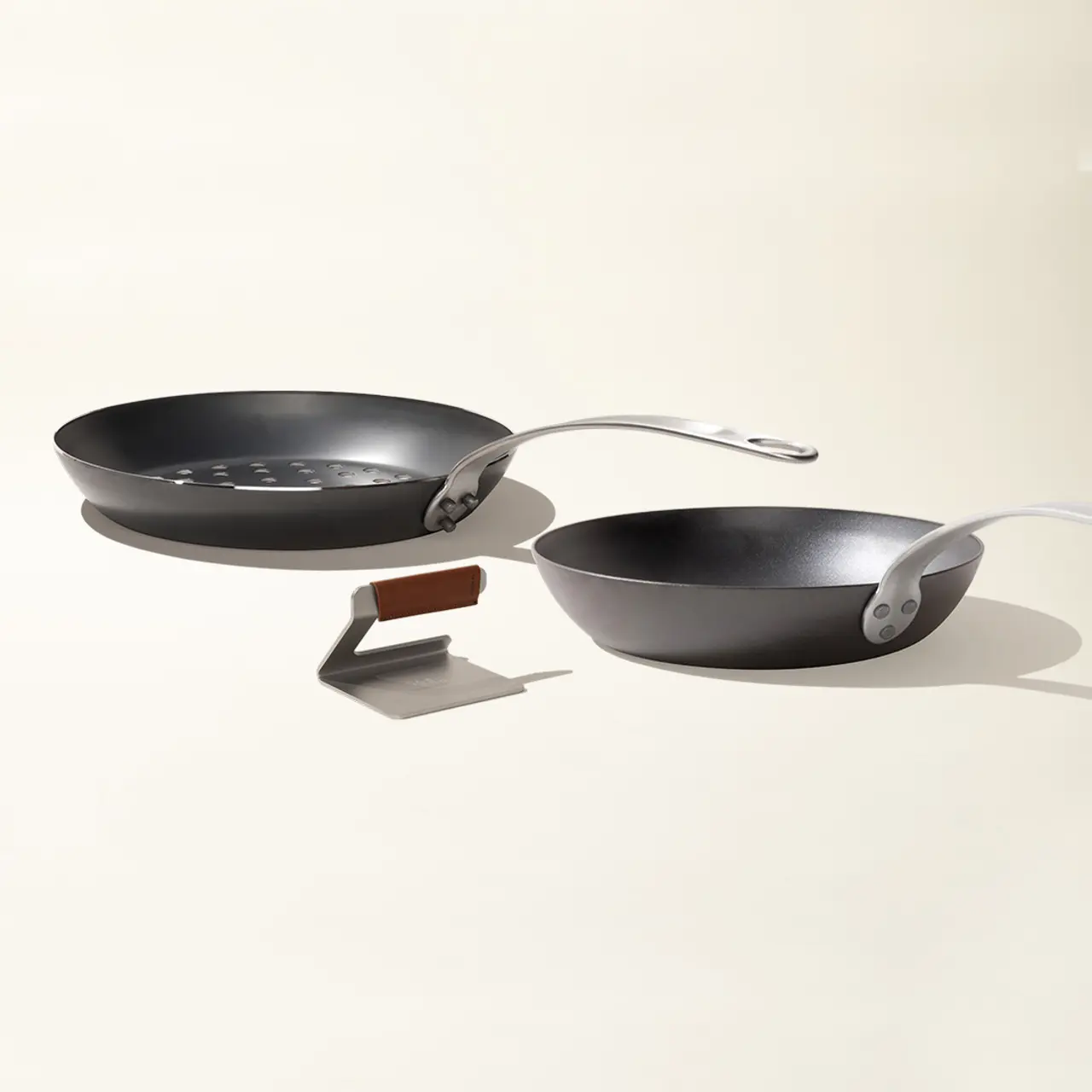 Two frying pans are balanced on a brown rectangular object, casting soft shadows on a light surface.