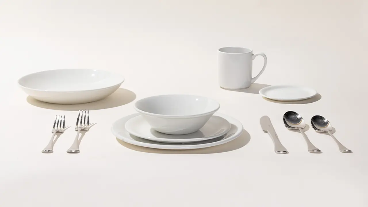 A neatly arranged set of white dinnerware accompanied by silverware on a plain background.