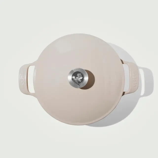 A round, cream-colored Dutch oven with a stainless steel knob sits against a plain background.