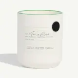 A white candle with a green rim features a label detailing its ingredients and burn time, including notes of pink rhubarb, Thai basil, violet leaf, fennel seed, and ginger root.