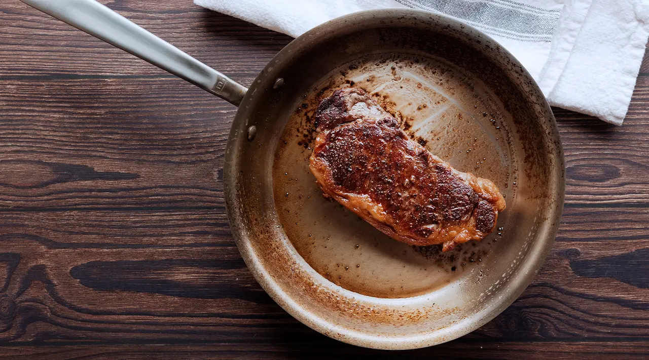 A succulent piece of steak is searing in a pan on a wooden surface.