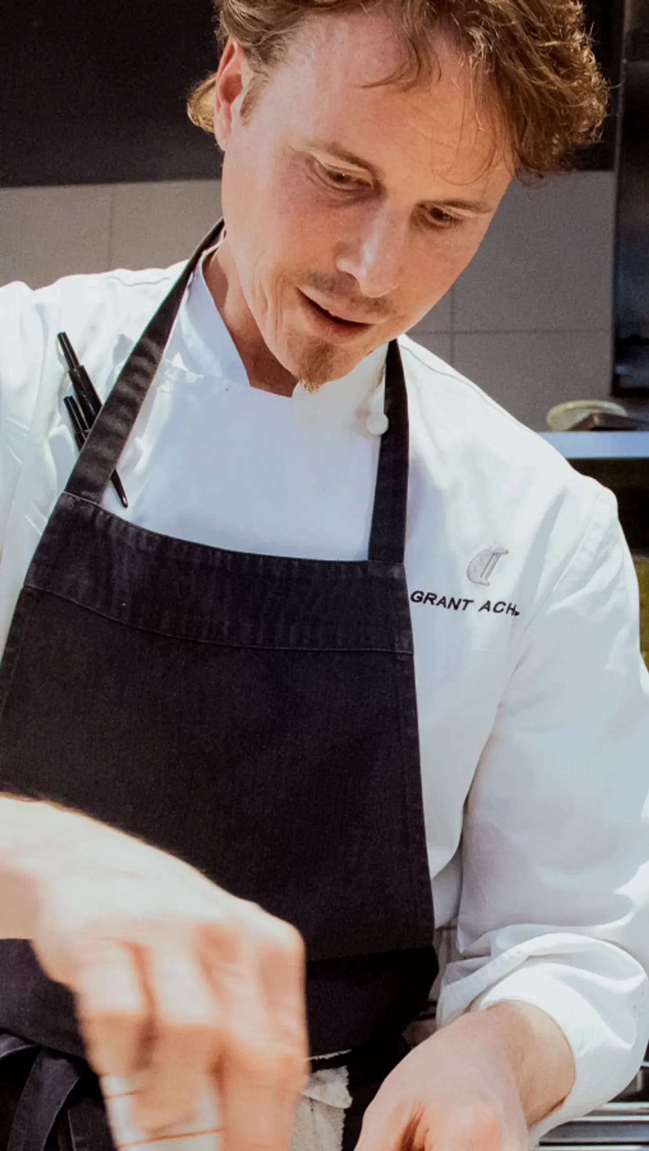 A chef in a white jacket and dark apron, with his name embroidered on it, is focused on preparing food.