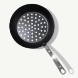 A black perforated pan with a silver handle lies against a light background.