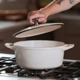 A hand is lifting the lid off a white cooking pot that is placed on a gas stove.