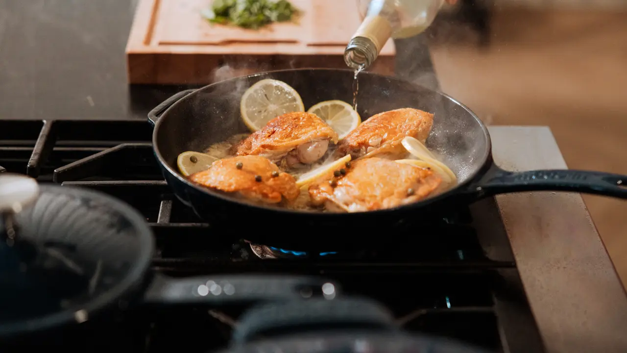 A skillet on a stove with chicken and lemon slices being deglazed with white wine.