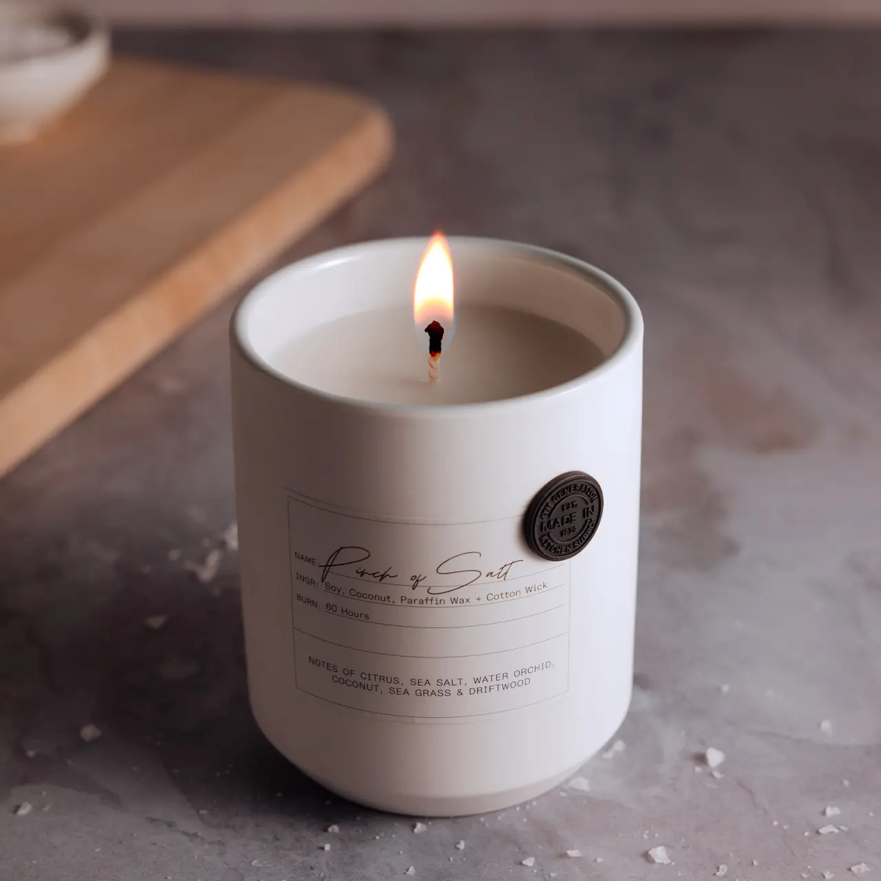 A lit scented candle with a tranquil flame and visible brand label rests on a muted surface.