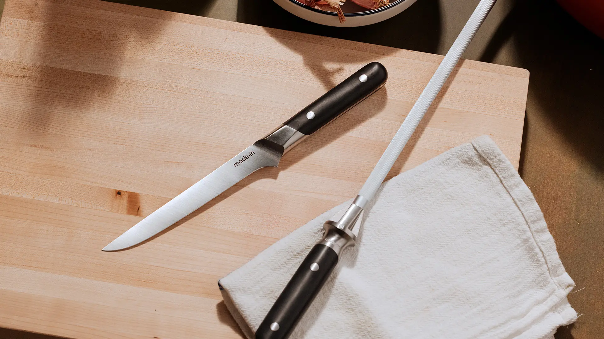 A knife and a honing steel lie on a wooden cutting board next to a folded white dishcloth.