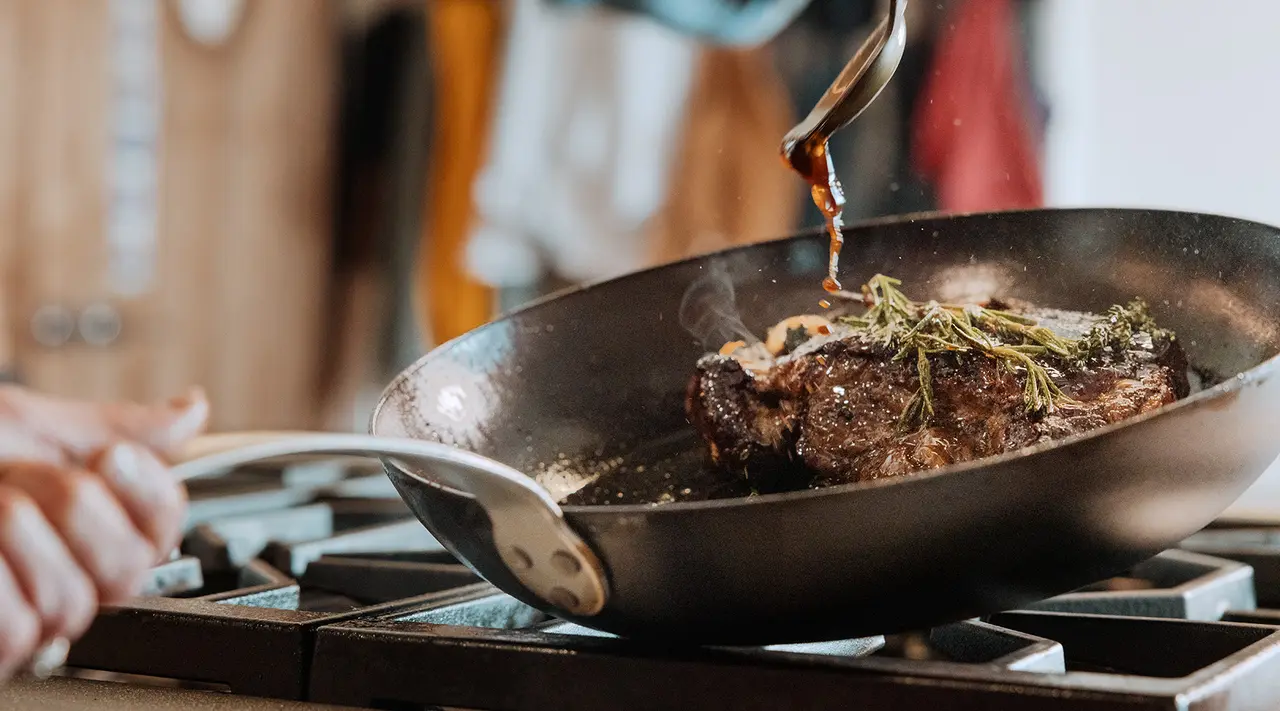 A juicy steak is being cooked in a frying pan with herbs on top, while someone's hand guides the pan on a gas stove.