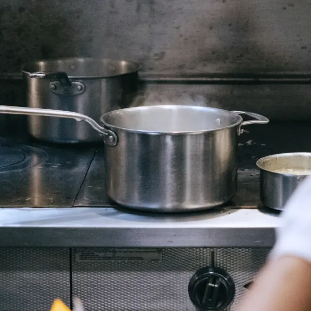 A stainless steel pot sits on a stovetop, emitting steam, with additional cooking pots in the background.
