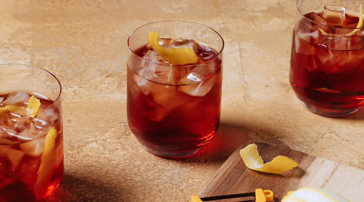 Three glasses filled with a amber-colored cocktail garnished with ice and lemon peels are set against a textured backdrop.