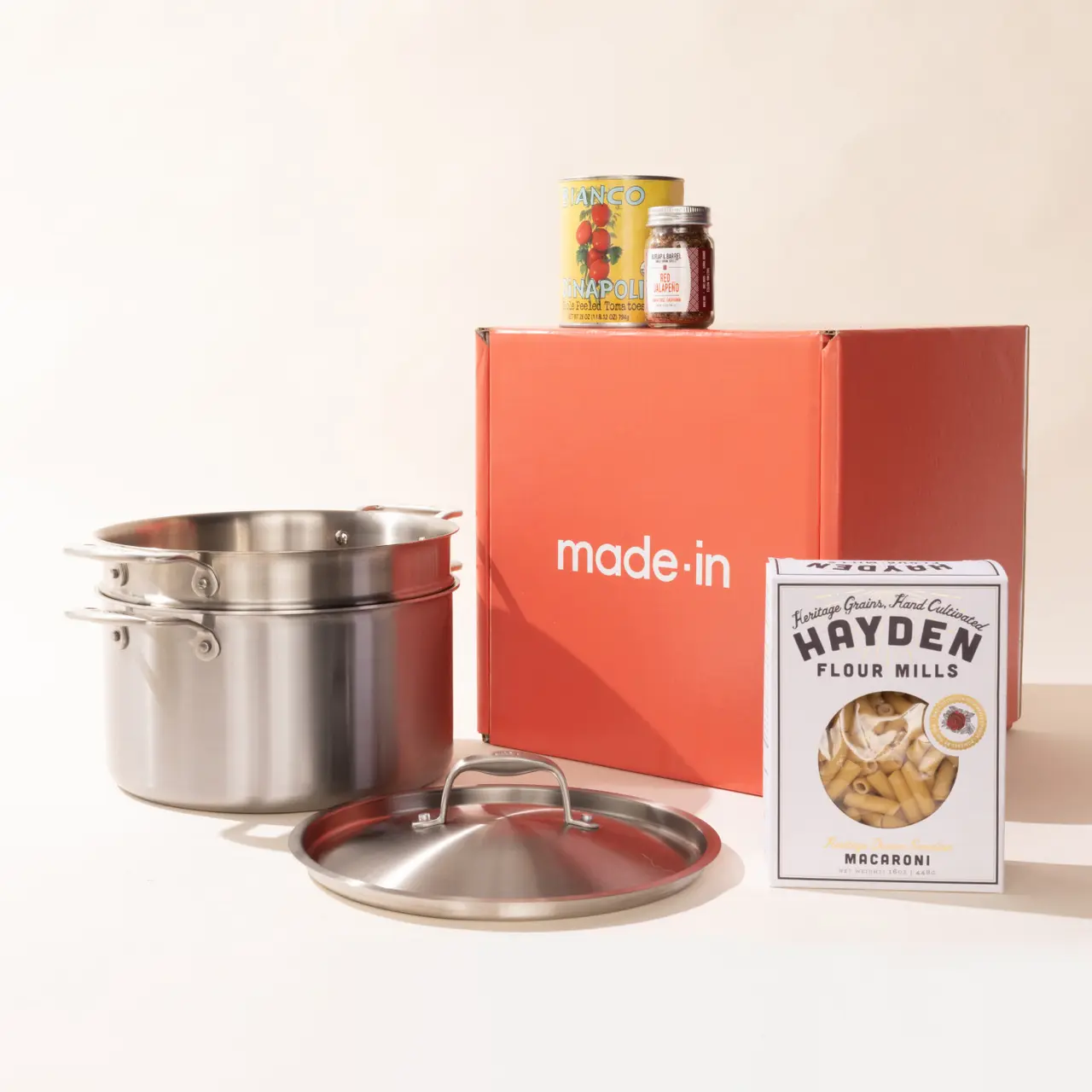 Cookware and food items including a pot, a can of tomatoes, and a box of macaroni beside a 'made-in' branded orange box on a neutral background.