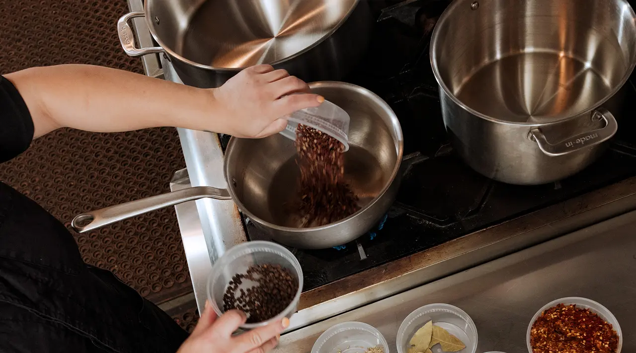 A person is pouring dry beans into a stainless steel pot from a clear measuring cup, with other ingredients and pots nearby on a kitchen counter.