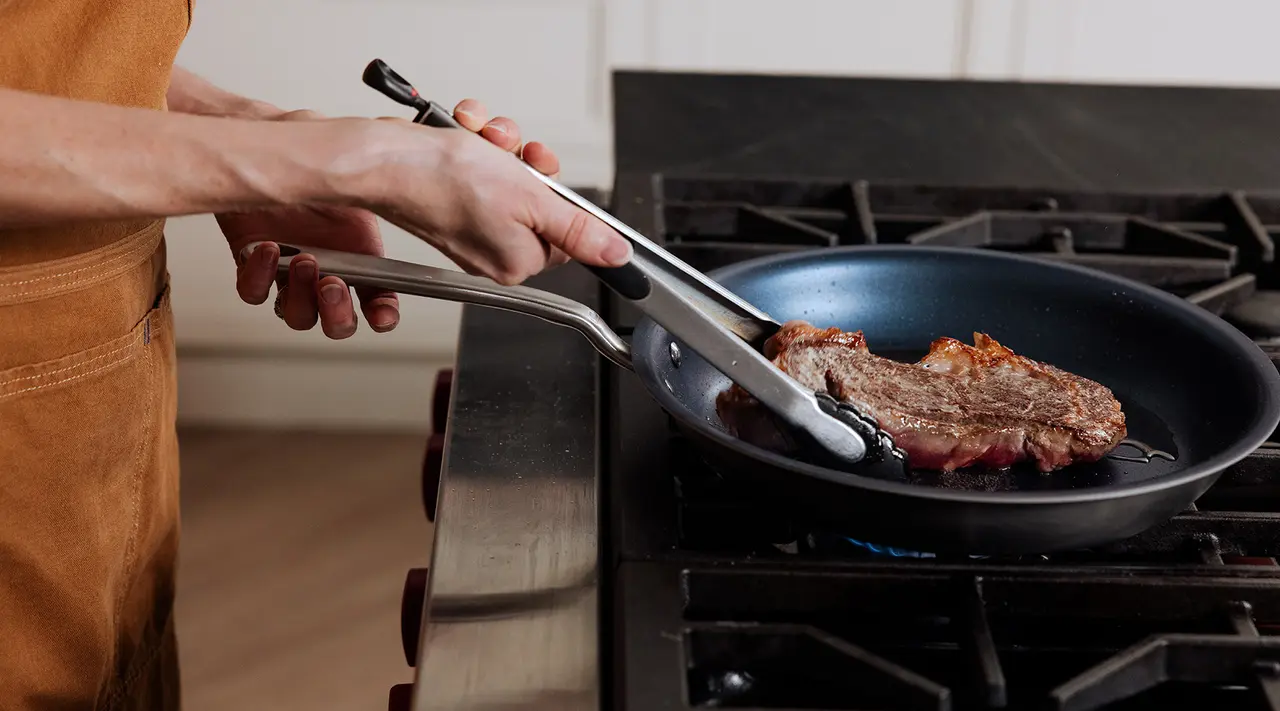 A person is cooking a steak in a skillet on a stove using tongs.
