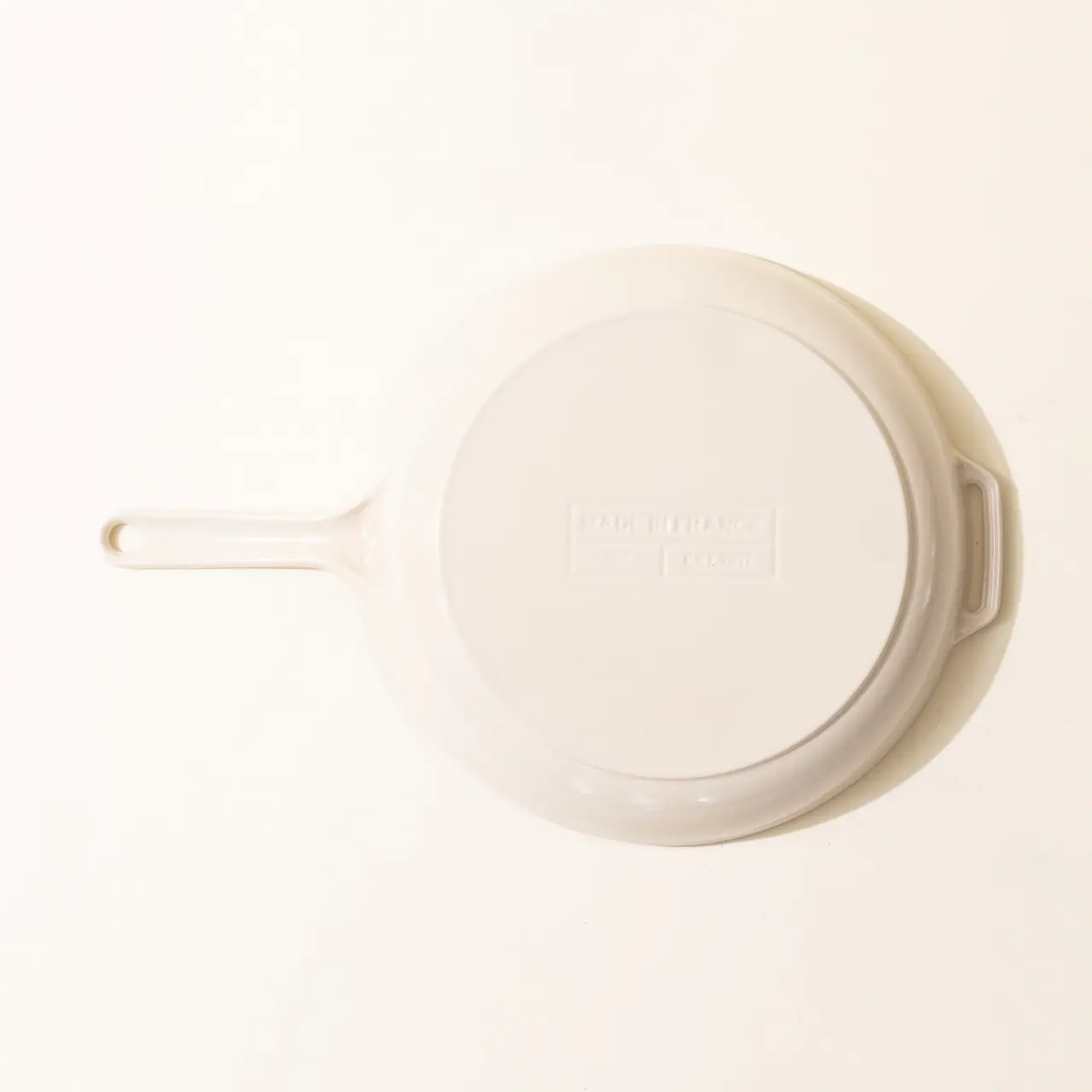 A white, ceramic skillet with a handle lies top-down on a light surface, displaying a text logo on its bottom.