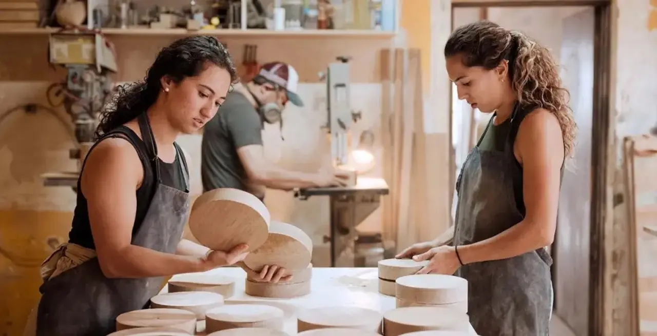 Two women in aprons are sanding wooden pieces in a workshop, with a man working in the background.