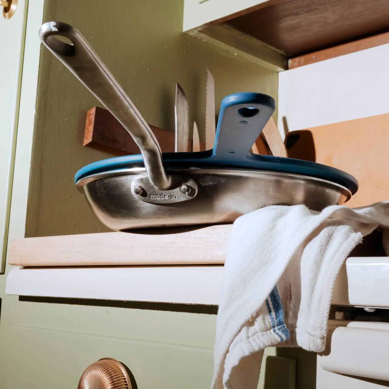 A stainless steel pan with a pair of tongs and a kitchen towel are placed on an open drawer in a home setting.