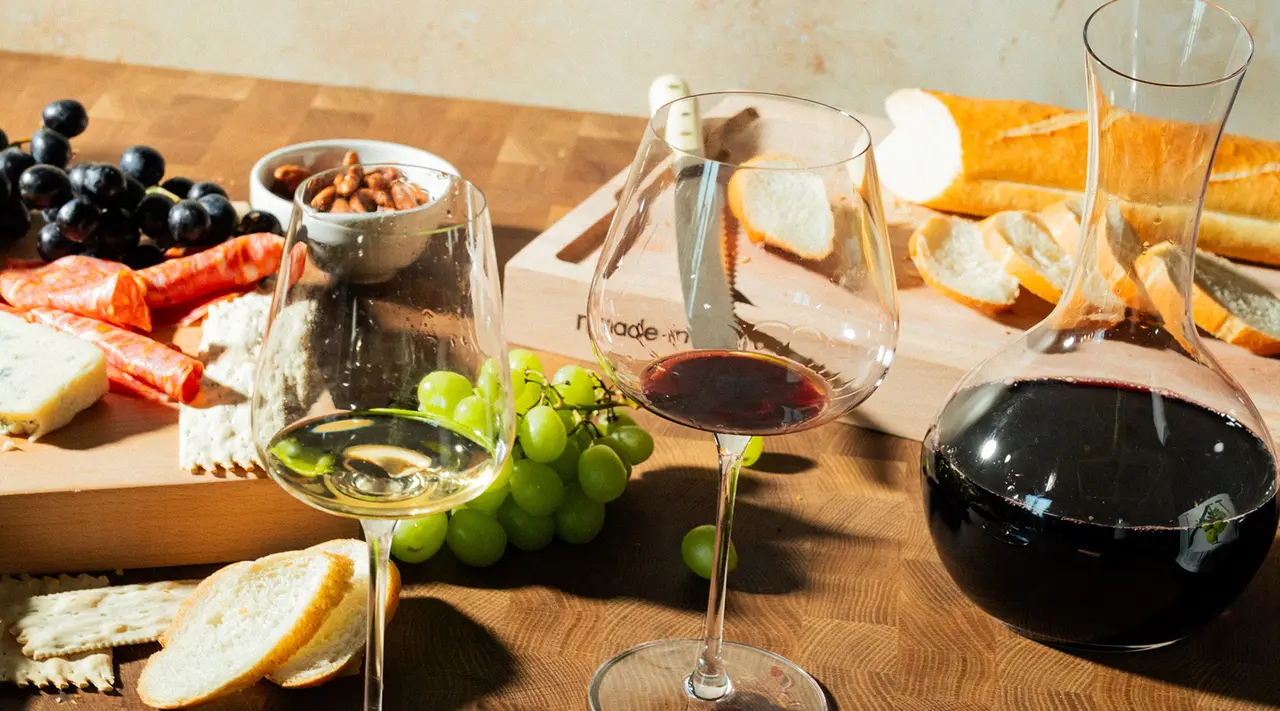 A spread of various cheeses, meats, nuts, grapes, bread, and two glasses of wine next to a wine decanter on a wooden surface.