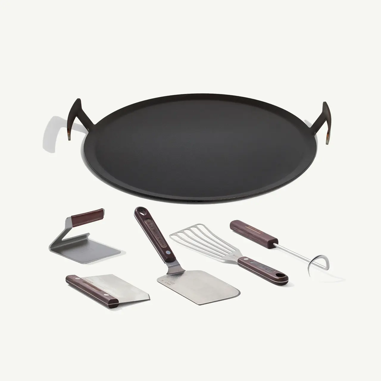 A black pizza pan is surrounded by various pizza-making utensils, including a cutter, spatula, and dough scraper, on a light background.